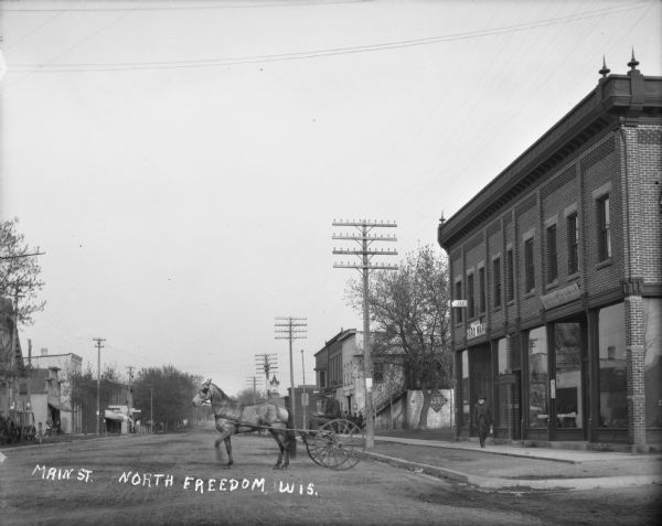 Man with horse and cart on Main Street. A brick commercial building on the right includes a hardware store and general merchandise store.