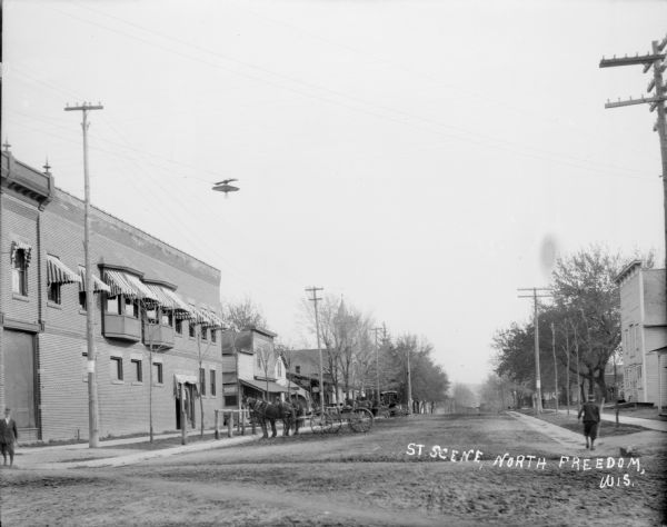 View down unpaved street. Horses pulling wagons are tied to posts at the curb. There are pedestrians along the sides of the street. Awnings are above windows of a two-story brick building on the left. There is a sign for shoes on a storefront further down the street. A streetlight is suspended on wires above the street.