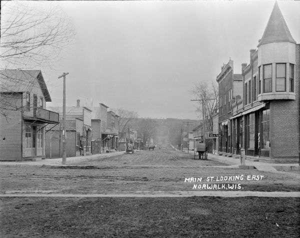 View down Main Street. There is a bank on the right and a 5&10 cent store on the left. Horse-drawn wagons and carriages are along the unpaved street. In the far background is a steep hill or bluff. On the sidewalk in front of the general store on the right is what appears to be a carbide/acetylene street light, with a glass bulb on top, and a tank on the bottom.