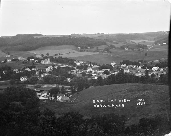 View of Norwalk looking down from a hill. Farm fields and forests surround the town.