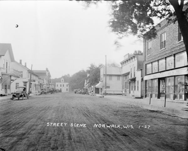 Street scene down middle of street, with businesses on both sides. A hotel, the post office, and a hardware store are on the left. A mercantile, ice cream shop and a gas station are on the right. Cars and trucks are parked along the curb on both sides. At the end of the street is a building with what appears to be a bell tower.