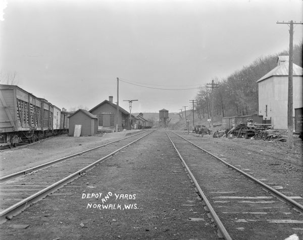 View down middle of two railroad tracks towards the railroad depot. Freight cars are on another set of railroad tracks on the left, and in the far background is a water tower. Men and women stand near the station, and three other men are near a platform on the right.