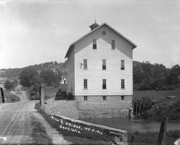 View across bridge of mill. There is a man standing on a wagon near the side of the mill. Behind the mill is a steep hill, with a house visible among trees on a rise just behind the mill.