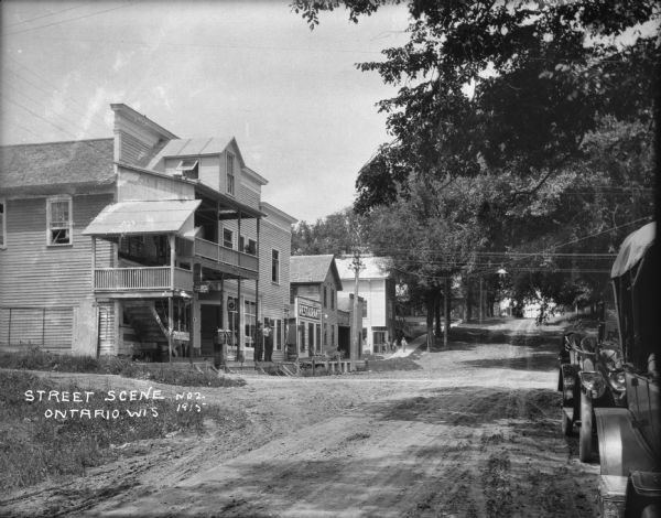 Street scene with a gas station and restaurant on the left and parked cars on the right. Two men stand in front of the service station, and further down the street people are in front of a building near a doorway with a sign that says: "Strickly Cash."