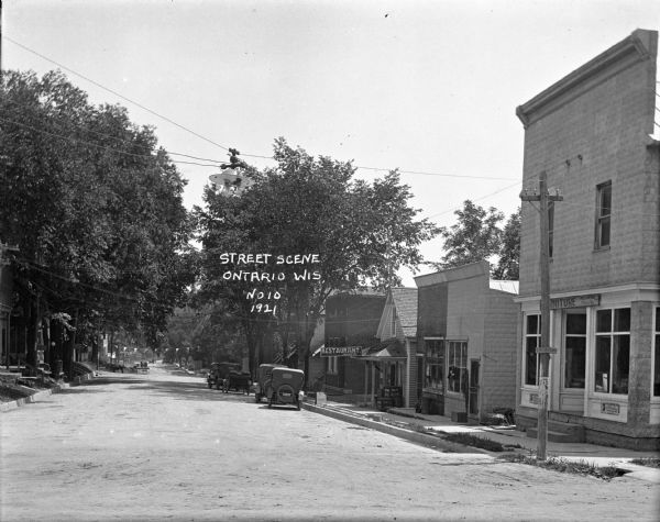 View down street with a restaurant and furniture store on the right. Automobiles are parked along the curb, and a street lamp is suspended from wires above the street. Horse-drawn wagons are in the street in the background.