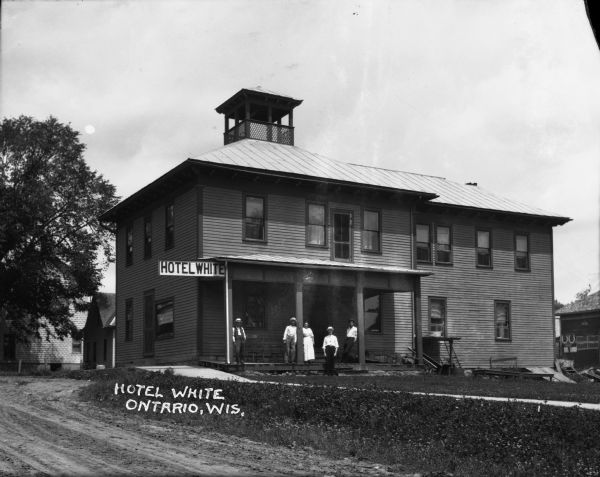 Exterior view of the Hotel White. Four men and one woman are posed on the porch. On the roof of the hotel is a widow's walk.
