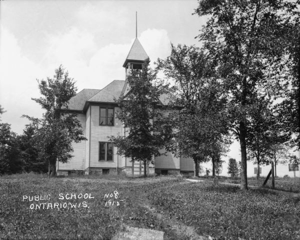 Exterior view of the public school building and grounds. There is a bell tower above the entrance, and trees are in the yard among tall grass.