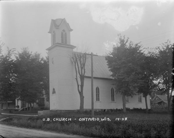 Exterior view of the United Brethren Church which has an arched window above the entrance and arched windows along the sides. Trees and homes surround the church. The steeple above the entrance has a belfry without a spire.