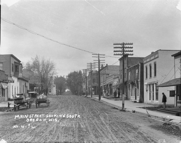 View looking down unpaved Main Street, with wagons and carts parked near the curb on the left near a building with a sign for a "Garage." Pedestrians are along the sidewalk on the right, in front of storefronts with awnings which are folded up. Businesses include a restaurant and ice cream shop, and a barber's pole is on the sidewalk.