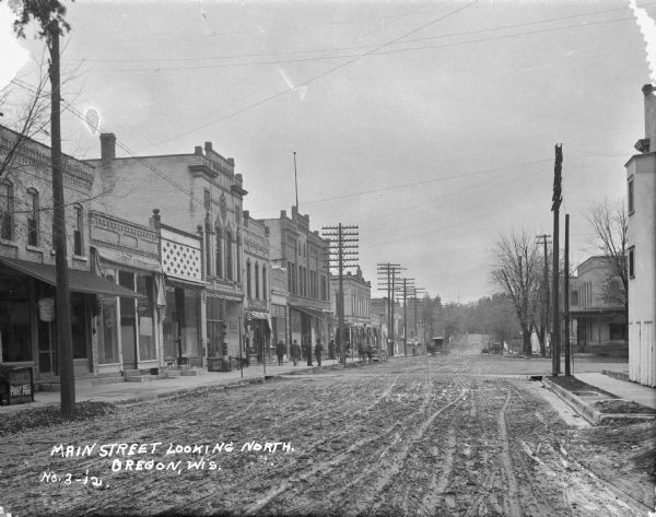 View down unpaved Main Street. There is a T-intersection on the right. On the left are a row of storefronts with a dog and pedestrians on the sidewalk. Businesses include a paint or hardware store. Most of the storefronts have awnings which are all pulled back. Horse-drawn carriages and carts are in the street in the background.