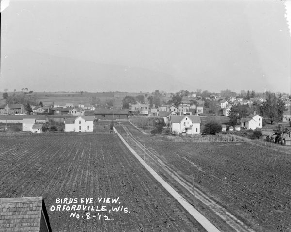 Elevated view of town with farm fields in the foreground, and low, rolling hills in the background. There is a church steeple in the far background on the right, and an awning on a building in the middle says "Shoes."