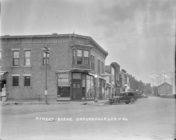 View across street of the Farmers Merchants Bank on a corner. On the right are parked cars along the curb in front of "Onsgard Bros." which has a Red Crown Gasoline gas pump on the sidewalk in front. Five lit streetlamps are along the sidewalk. There is a sign above the sidewalk further down the street for the "I.O.O.F."