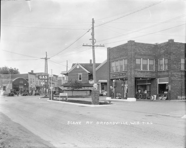 View across street towards the Honeysett Oil Co. Filling Station, which is a two-story brick building. A woman and two children are on the stoop of a house behind the gas station. Further down the street, which curves towards the right, is an ice cream shop, and the Hotel Orford. More businesses are on the left side of the street in the background.