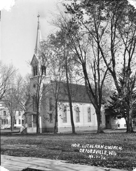 Exterior view of the Norwegian Lutheran Church with steeple. The church has arched stained glass windows in the front and on the sides, and is surrounded by a lawn and trees. A large house is in the background on the left.
