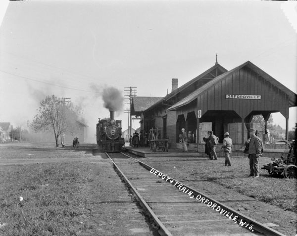 View down railroad tracks towards depot with railroad train pulling into the station. Passengers are waiting on the platform, and other people are standing near carts in the right foreground.