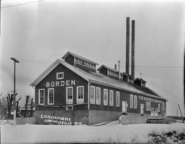 Exterior view of the Borden Condensery and grounds. Snow covers the ground. The right side of the building has two doors elevated above the driveway for a loading dock. Two tall chimneys rise out of the center of the roof, which also has four large roof vents with windows.