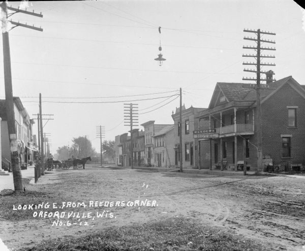 Street scene looking east from the corner of the Reeder's Hotel. Men stand near horses and wagons on the left near a sidewalk and storefronts. A small dog stands near one of the horses. A streetlamp is suspended from wires above the street.
