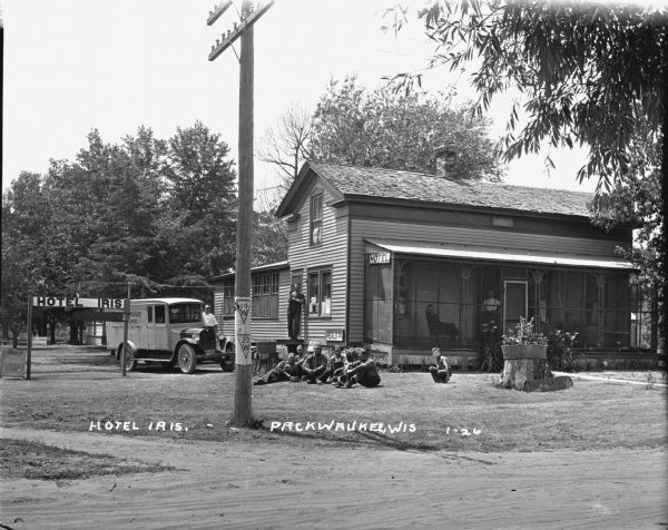 View across road of the Hotel Iris, with a large group of men and boys posed in the yard, on the screened-in porch, and near a delivery van parked on the left.