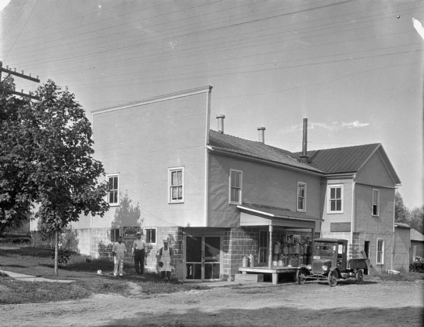 View across road of creamery building. Male workers are standing near the entrance on the left in front of a sign that says "Patch Grove Creamery." On the right a delivery truck is parked near the loading dock. The dock has a roof, and milk cans are stacked underneath.