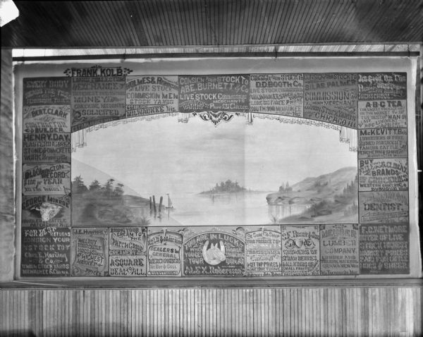 A stage curtain with advertising for local businesses, and a landscape scene in the middle depicting a sailboat near a shoreline with an island in the distance. The name "Frank Kolb" is painted on the top left of the curtain.