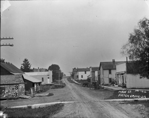 View down Main Street. On the left is a barn covered with circus broadsides. Across the street is a building with a storefront and awning, and a sign that says: "Meals and Lodging at Mrs. England's." Carriages and carts are parked along the curb in front.