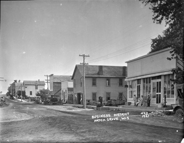 View across street towards the row of businesses along the sidewalk on the right. Includes, from the right, a man in an apron, and three young girls standing in front of a general store and post office. Further down the street is a two-story building with two wooden wheels in front of a large, open double doorway. There are a few more commercial buildings, and then dwellings further down the street. There are various signs posted for "Shoes," "Mobiloil," and "Ice Cream." Automobiles are parked along the curb.