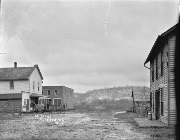 View down street with three or four commercial buildings. On the right is a sign for a telephone pay station on a two-story clapboard building, and behind it is a one-story building further. Across the street on the left a group of man and horses with a wagon stand near two more commercial buildings. There is a woman standing in the window on the second floor of the building on the left. Snow is on the ground, and in the distance are more scattered wooden buildings in a field, with a tree-covered hill in the far background.