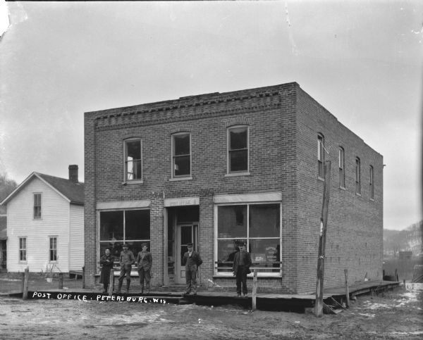 View across street of a group of men standing on the wooden sidewalk in front of the post office/general store. Above the door is a sign for the "Post Office." There is a house on the left. Snow is on the ground and in the far background is a hill.