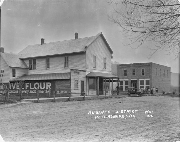 View across street of commercial buildings, with a tree-lined hill in the far background. A group of men and women are standing on the porch of the brick building along another street on the right. One of the men is holding a baby. A car is parked near them at the curb. On the other side of the street a man stands on the porch of the post office. On the left side of the building is an large advertisement for "MARVEL FLOUR".