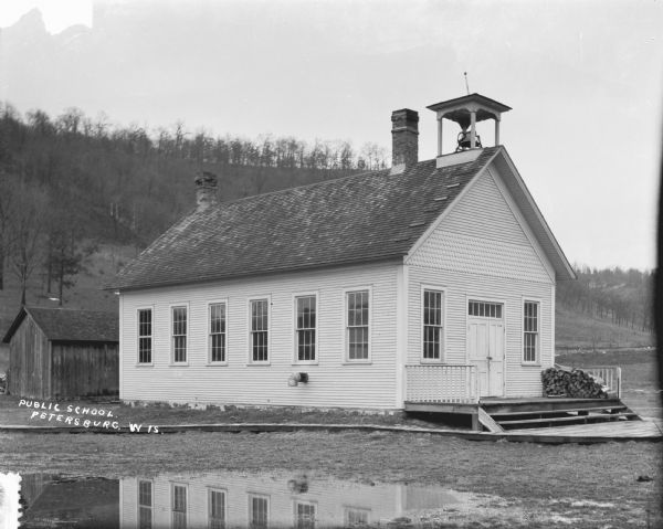 View of the public school with two chimneys, a bell tower, and firewood stacked on the porch. The building is reflected in the puddle in the foreground. Behind the school is a small outbuilding, and a tree-lined hill rises in the far background.