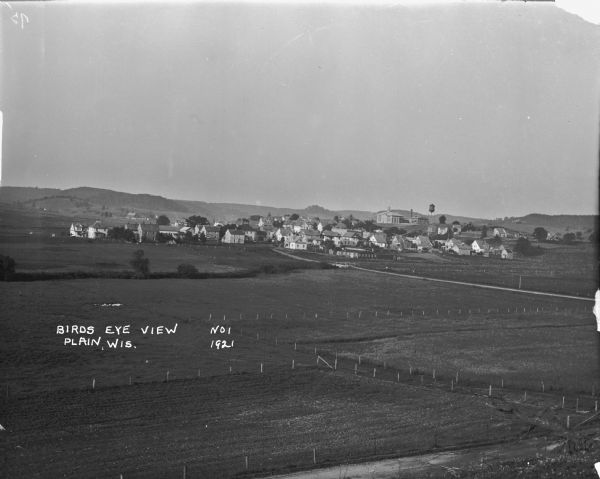 Elevated view over farm fields in the foreground and town in the distance, with low rolling hills in the far background. Buildings that stand out are St Luke's School, and a water tower.