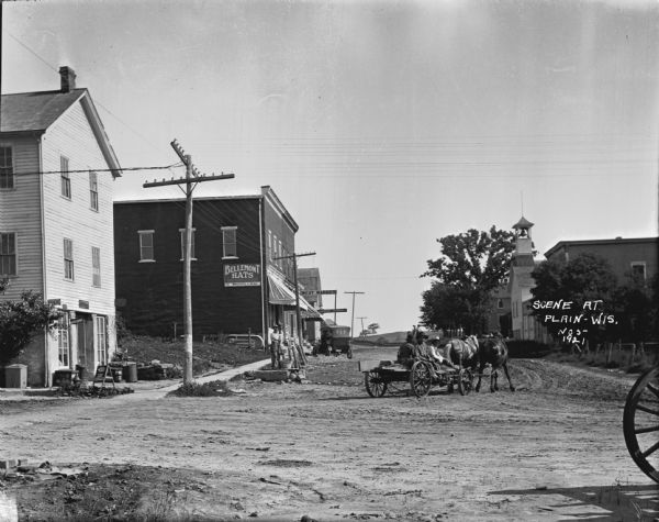 View of men standing near a T-intersection. The three-story building on the left side of the intersection has a sandwich board advertising "Ice Cream." A man stands near a hand-pump near the side of the street, and  three men sit on a horse-drawn cart. Further down the street is a hat shop and meat market on the left, and a school with a bell tower on the right.