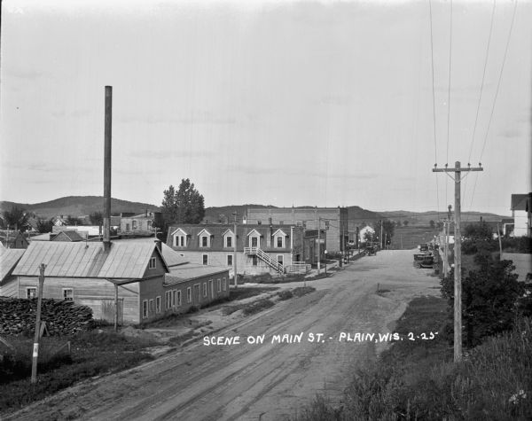 Elevated view of Main Street, with industrial and commercial buildings. Cars are parked at the curb towards the far end of the street, and fields and tree-lined hills are in the distance. There is a large stack of firewood near the building in the foreground on the left.