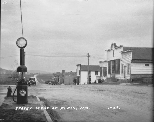 View from town of the road leading out of town towards fields and low hills. On the left at the curb is a fire hydrant and a Red Crown Gasoline gas pump. Automobiles are parked along both sides of the road in the background near brick commercial buildings, including a confectioner/meat market and a garage. The storefront across the street also has a Red Crown Gasoline gas pump.