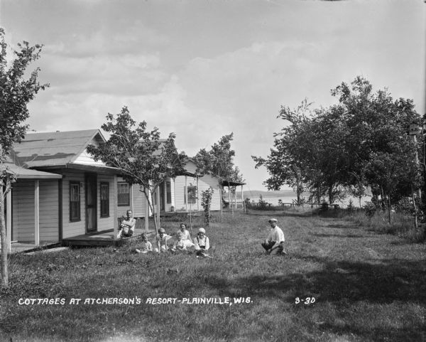 View of resort cottages near a lake. A group of vacationers are sitting on the lawn and porch. A birdhouse on a tall post is among trees on the right. Hills are in the distance on the far side of lake.