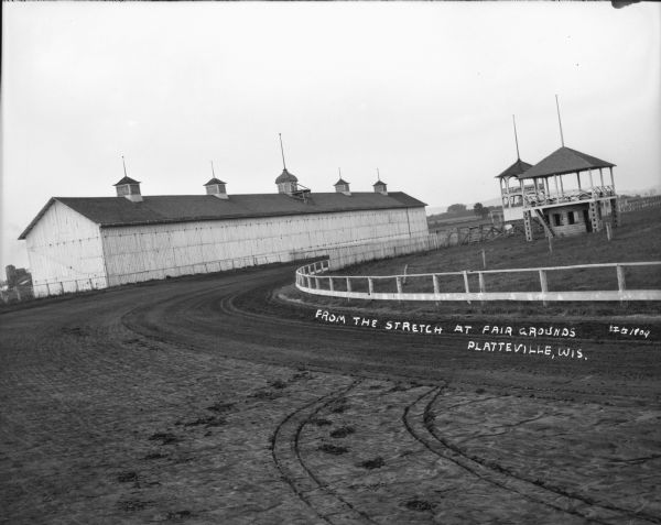 View across fairground racetrack, with judging stands and large barn in the background.