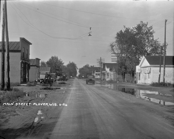 View of Main Street with puddles along the side of the street. Trucks and automobiles are traveling down the street, and are parked on the left. A restaurant and hotel are on the left and a meat market and soda shop are on the right.