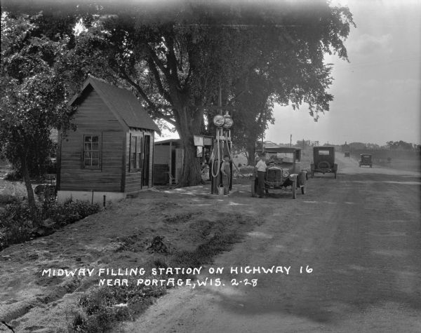 View down road towards a filling station near Portage. A man stands while filling his tank. Two boys stand nearby. Under large trees are two pumps, a small building and a vendor's booth. Three automobiles are traveling down the road in the far background.