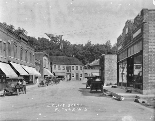 View down street of the central business district. A large brick building on the left with awnings includes a furniture store. Automobiles are parked at an angle along the curb, and pedestrians are along the street. On the right is a storefront, with a dog on the sidewalk in front. Behind is a garage, and in the background is a post office. A flag and banners are suspended over the street on wires.