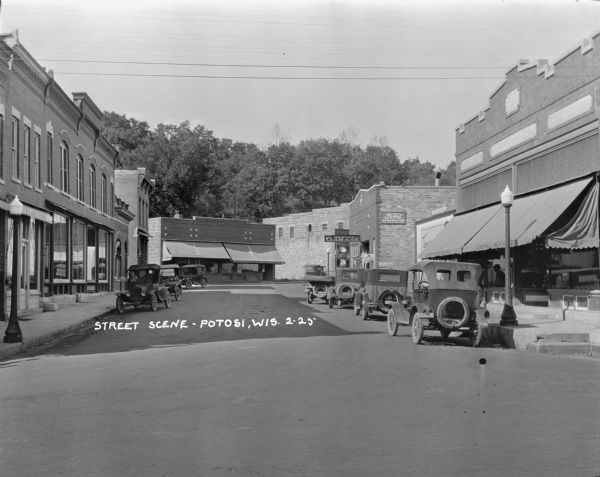 View down street of the central business district. There is a furniture store on the left, a general store and service garage on the right and a drugstore in the background. Automobiles are parked in the street along the curb.