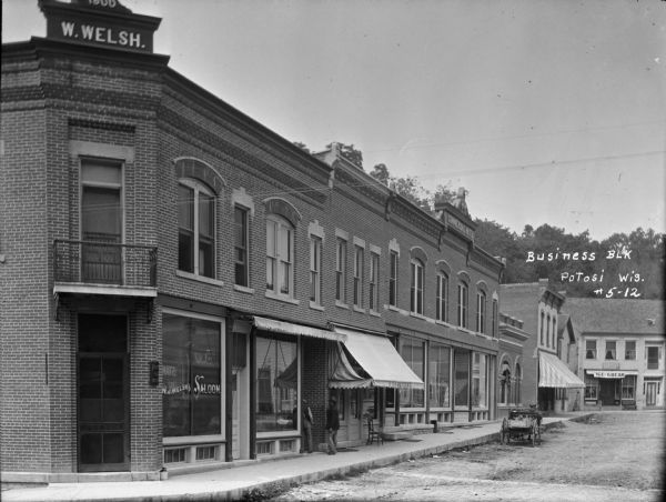 View from street towards a row of brick buildings. On the left corner is a building with a sign on the roof that says "W. Welsh." The entrance has a balcony above it, and the large window says "Saloon." A furniture store, dentist office, Commercial Hotel, and ice cream parlor are along the sidewalk in the background. Two men are standing in a doorway. A horse-drawn cart is parked in the street along the curb.