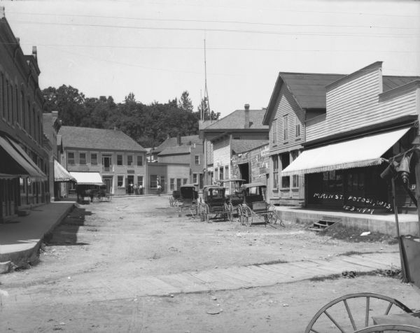 View down Main Street. On the right is a general store, and Livery/Hardware building with horse-drawn carts parked in the street in front. A group of men are standing in front of the post office, dentist office and barbershop buildings in the background. A horse-drawn cart is parked in front of the carpet store on the left.