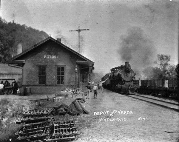 View towards depot of a train pulling up to the platform. A group of men are on the far left behind the depot. Metal parts, firewood and wooden crates are stacked in the foreground. A group of men and boys are standing on the platform.