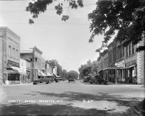 View down street at intersection of commercial buildings on both sides. On the left is a drug store/soda fountain, restaurant and a cafe. On the right is a real estate/insurance office, a theater, and a general store. A car is traveling down and the street, and other cars are parked at an angle along the curbs.