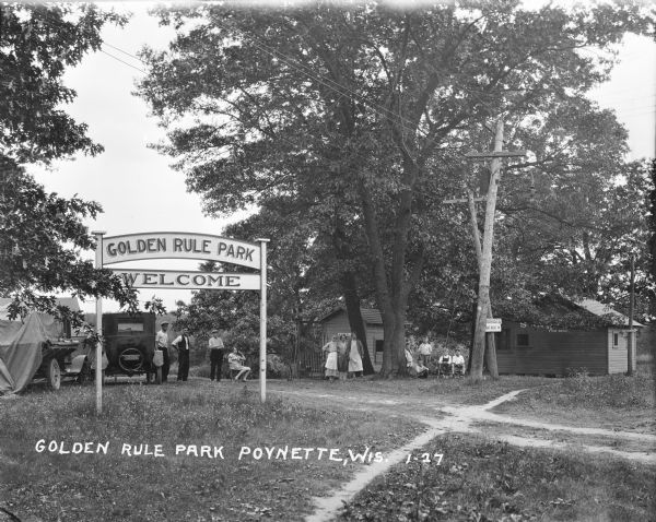 View across lawn towards the entrance to Golden Rule Park. A large sign says "Golden Rule Park / Welcome." Men, woman and children are posed near cabins and automobiles. One of the automobiles is covered with canvas.