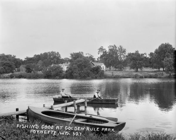 View from shoreline of a pair of men fishing in a lake. Another rowboat is tied at a pier in the foreground. On the far shoreline are cottages and trees.