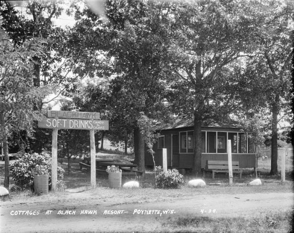 View across road of cottage with a screened porch in a wooded area. Picnic tables are on the left. A bench is in front of the cottage. A row of planters in the foreground are flanking a large sign advertising soft drinks, candy and cigars.