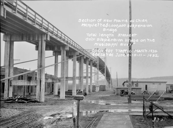 "Section of New Prairie du Chien Marquette $1,000,000 Suspension Bridge. Total length 3729 feet. Only suspension bridge on the Mississippi River. Open for traffic March 1932. Dedicated June 9-10-11 1932."<p>View from below of a section of the Marquette Suspension Bridge. There is a mailbox in the foreground near a sidewalk and a wire fence. On the left is a stone building behind the bridge supports. The road, which is muddy and has puddles, leads to freight cars sitting under the bridge in the background. An automobile is parked nbear a contractor's on the right in the background. In the far distance is a hill with a ridge.