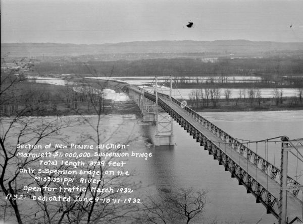 "Section of New Prairie du Chien Marquette $1,000,000 Suspension Bridge. Total length 3729 feet. Only suspension bridge on the Mississippi River. Open for traffic March 1932. Dedicated June 9-10-11 1932."<p>View from above of the Marquette Suspension Bridge over the Mississippi River and wetlands. A town and rolling hills are in the background.