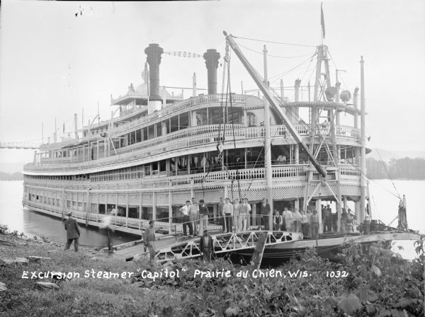 View from shoreline of the "Capitol" excursion steamer pulled up to the bank of the Mississippi. The steamer has three or four decks, and elaborately detailed railings and ornamentation. A crane attached to the front is attached to a attached to a passenger boarding bridge. Passengers and workers are gathered on the decks and on the boarding bridge. The Marquette Suspension Bridge in the far background.